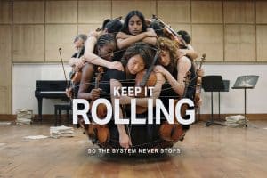 Eine Gruppe junger Musiker:innen mit Geigen und Bratschen ist in einem Proberaum eng ineinander verschlungen und bildet eine kugelförmige Komposition. Im Vordergrund steht der Schriftzug „KEEP IT ROLLING“, darunter „SO THE SYSTEM NEVER STOPS“.