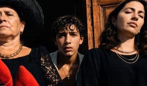 Nahaufnahme von drei Menschen vor einem dunklen Hauseingang in grellem Sonnenlicht. Links eine ältere Frau mit schwarzem Hut und Spitzenkleid, in der Mitte ein junger Mann mit feuchten Haaren und Kreuzkette, rechts eine Frau mit geschlossenen Augen und goldenem Schmuck. Starke Hell-Dunkel-Kontraste verleihen der Szene eine dramatische Atmosphäre.