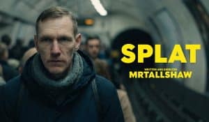 Filmplakat mit einem ernst blickenden Mann in dunkler Jacke und grauem Schal auf einem belebten U-Bahnsteig. Im Hintergrund verschwimmen wartende Menschen und ein Tunnel. Rechts steht in leuchtend gelber Schrift der Titel „SPLAT“, darunter „Written and Directed MRTALLSHAW“.