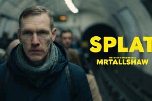 Filmplakat mit einem ernst blickenden Mann in dunkler Jacke und grauem Schal auf einem belebten U-Bahnsteig. Im Hintergrund verschwimmen wartende Menschen und ein Tunnel. Rechts steht in leuchtend gelber Schrift der Titel „SPLAT“, darunter „Written and Directed MRTALLSHAW“.
