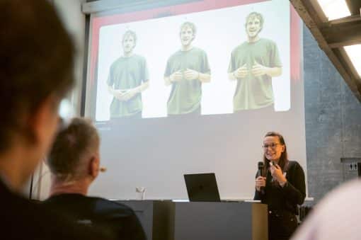 Frau auf der Bühne präsentiert, Gäste im Vordergrund, hinten eine Leinwand