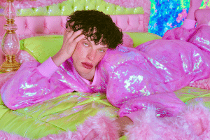 Ein junger Mensch mit dunklen Locken liegt gelangweilt auf einem neongrünen Bett, den Kopf auf eine Hand gestützt. Er trägt einen schillernden, pinkfarbenen Plüschanzug. Die überzeichnete Szenerie in leuchtendem Pink, Neongrün und Türkis vermittelt eine künstliche, poppige Ästhetik.