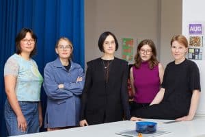 Gruppenporträt von fünf Frauen hinter einer weißen Theke mit hellgelben Schubladen. Links hängt ein kräftig blauer Vorhang, dahinter liegt ein heller Studioraum mit Strahlern und Plakaten an der Wand. Die Frauen tragen unter anderem ein hellblau gemustertes Shirt, eine blaue Jacke, eine schwarze Weste, ein magentafarbenes Top und ein schwarzes T-Shirt. Auf der Theke stehen blaue Glasschalen.