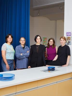 Gruppenporträt von fünf Frauen hinter einer weißen Theke mit hellgelben Schubladen. Links hängt ein kräftig blauer Vorhang, dahinter liegt ein heller Studioraum mit Strahlern und Plakaten an der Wand. Die Frauen tragen unter anderem ein hellblau gemustertes Shirt, eine blaue Jacke, eine schwarze Weste, ein magentafarbenes Top und ein schwarzes T-Shirt. Auf der Theke stehen blaue Glasschalen.