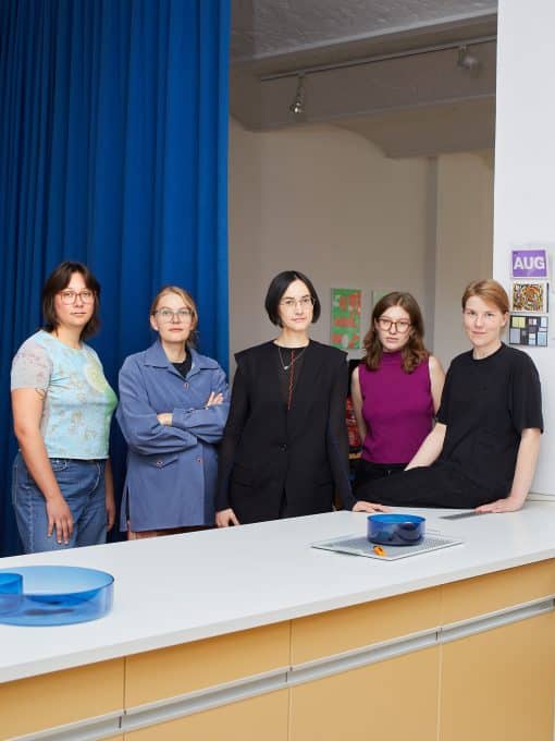 Gruppenporträt von fünf Frauen hinter einer weißen Theke mit hellgelben Schubladen. Links hängt ein kräftig blauer Vorhang, dahinter liegt ein heller Studioraum mit Strahlern und Plakaten an der Wand. Die Frauen tragen unter anderem ein hellblau gemustertes Shirt, eine blaue Jacke, eine schwarze Weste, ein magentafarbenes Top und ein schwarzes T-Shirt. Auf der Theke stehen blaue Glasschalen.