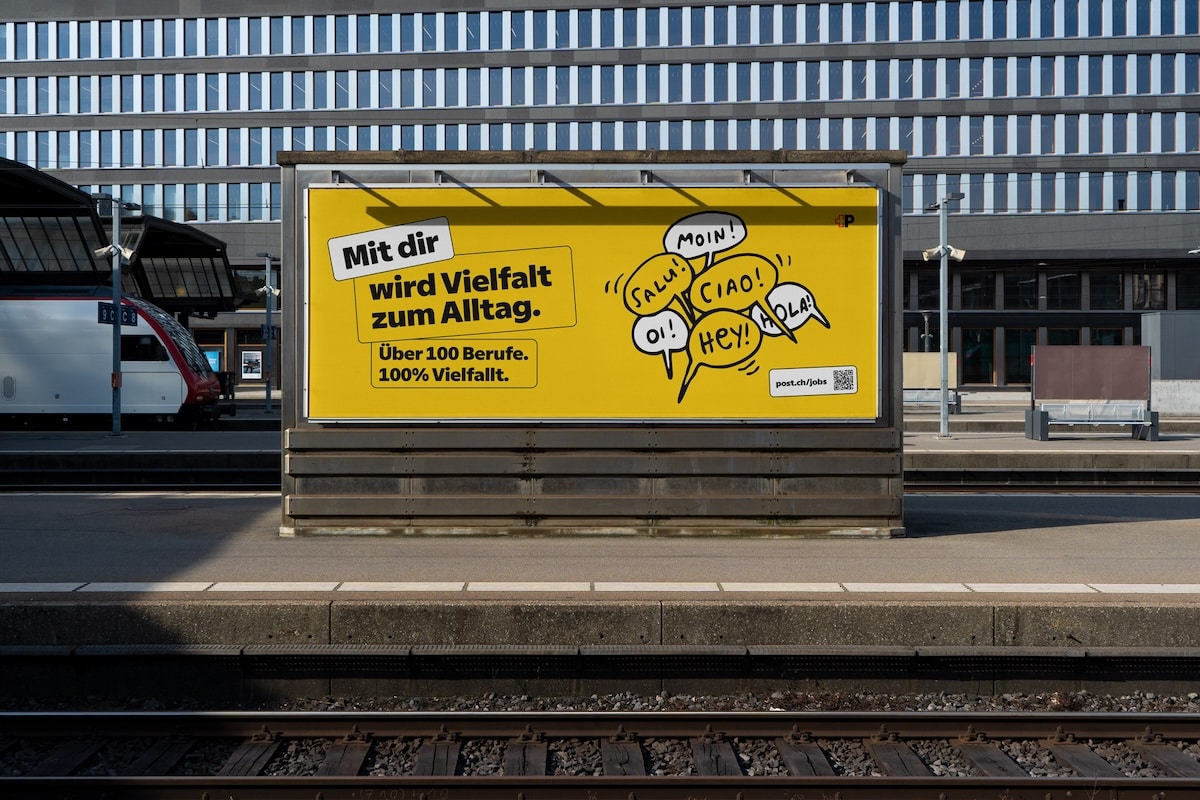 Ein gelbes Billboard der Schweizerischen Post mit dem Slogan „Mit dir wird Vielfalt zum Alltag.“ sowie gezeichneten Sprechblasen mit internationalen Grüßen wie „Moin!“, „Ciao!“ und „Salut!“.
