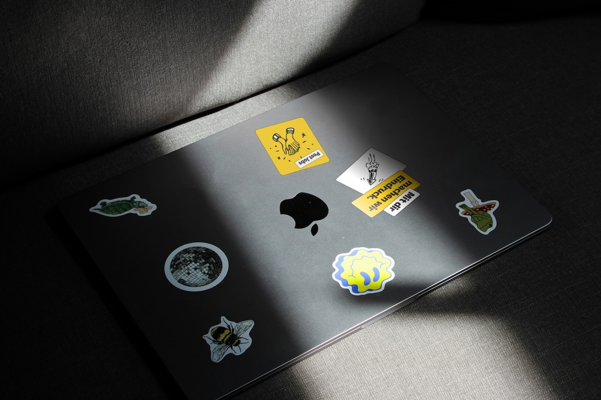 Ein geschlossenes, graues MacBook im Sonnenlicht, verziert mit bunten Stickern, darunter ein quadratischer gelber Post-Jobs-Sticker und ein Spruch „Mit dir machen wir Eindruck.“.