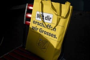Eine gelbe Baumwolltragetasche mit dem Aufdruck „Mit dir erschaffen wir Grosses.“ und einer kleinen Grafik lehnt im Halbschatten auf einem roten Klappstuhl.
