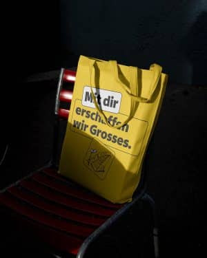 Eine gelbe Baumwolltragetasche mit dem Aufdruck „Mit dir erschaffen wir Grosses.“ und einer kleinen Grafik lehnt im Halbschatten auf einem roten Klappstuhl.