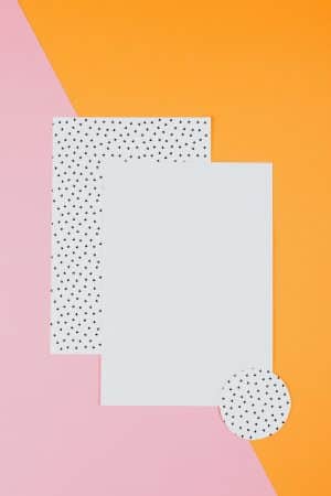 Ein weißes und ein mit schwarzen Punkten markiertes Blätter Papier, vor einem organge- und pinkfarbenen Hintergrund, Stil ist poppig-heiter
